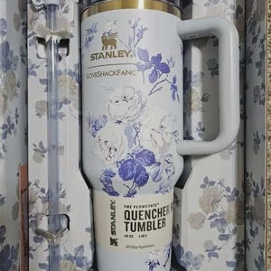 May include: A white Stanley Quencher tumbler with a floral design in a gift box. The tumbler has a handle and a straw. The floral pattern includes white roses and purple accents. The tumbler is 40 oz (1.18 L).