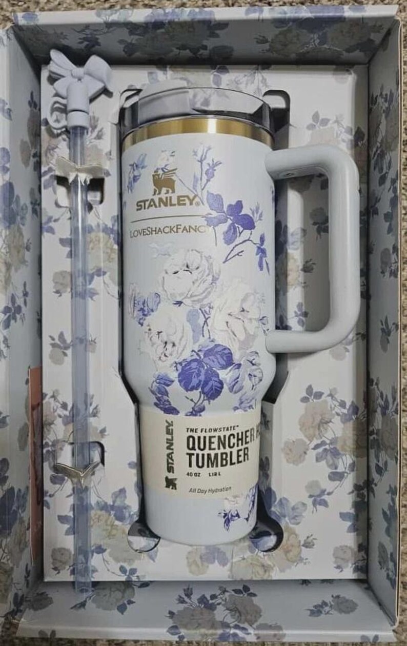 May include: A white Stanley Quencher tumbler with a floral design in a gift box. The tumbler has a handle and a straw. The floral pattern includes white roses and purple accents. The tumbler is 40 oz (1.18 L).
