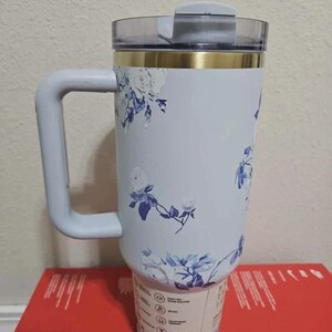 May include: A light blue insulated tumbler with a handle, featuring a floral design in blue and white. The tumbler has a clear lid and a gold band near the top. The handle is light blue.