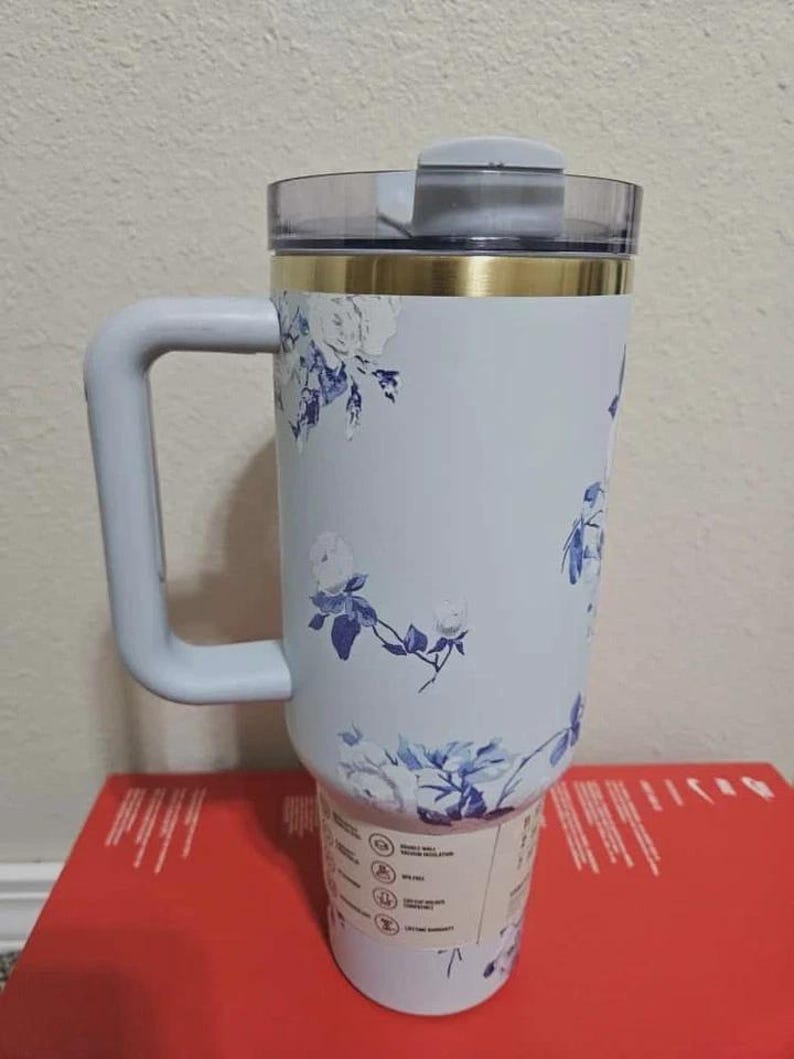 May include: A light blue insulated tumbler with a handle, featuring a floral design in blue and white. The tumbler has a clear lid and a gold band near the top. The handle is light blue.
