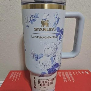 May include: A light blue Stanley Quencher tumbler with a floral design in white and blue. The tumbler has a handle and a clear lid. The text "STANLEY" and "LOVESHACKFANCY" are visible on the tumbler. The tumbler is 40 oz.