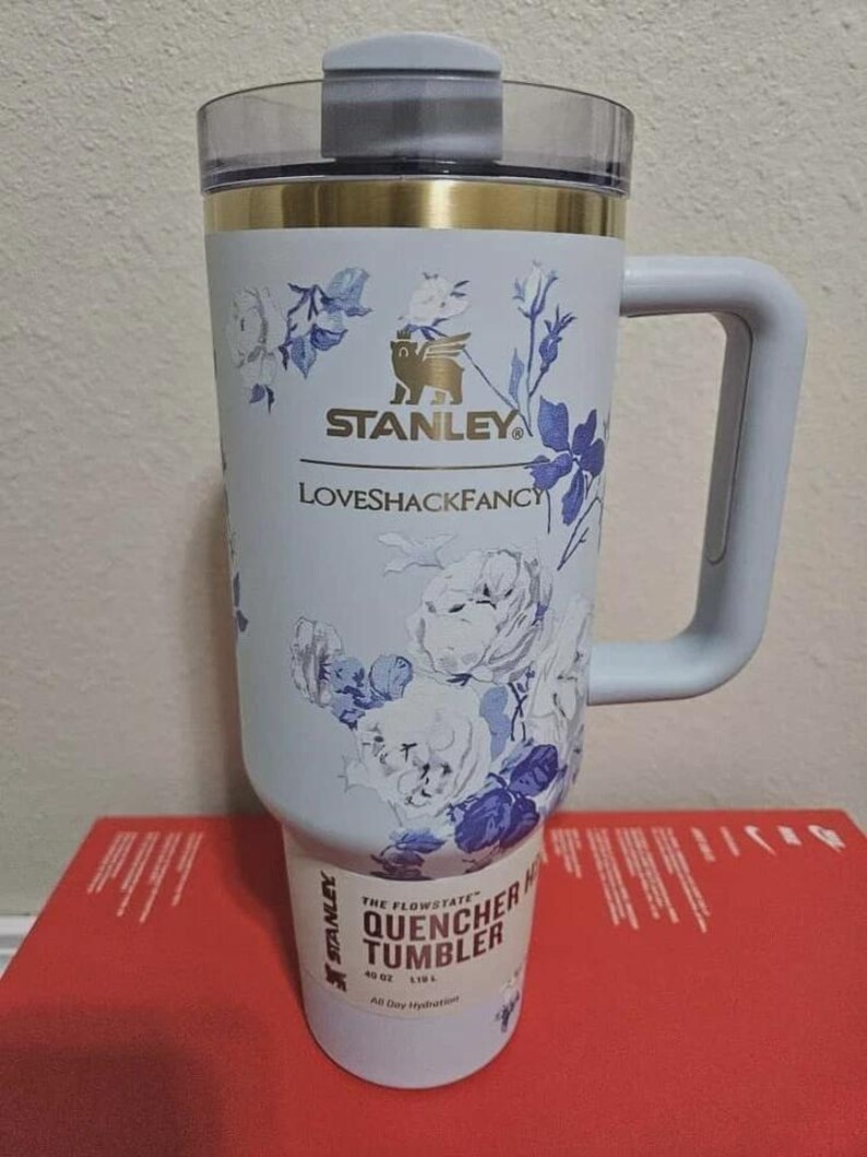 May include: A light blue Stanley Quencher tumbler with a floral design in white and blue. The tumbler has a handle and a clear lid. The text "STANLEY" and "LOVESHACKFANCY" are visible on the tumbler. The tumbler is 40 oz.