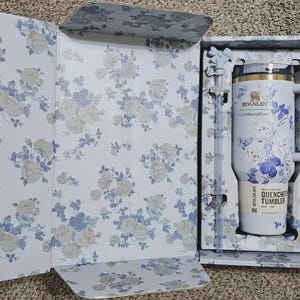 May include: A white Stanley Quencher tumbler with a handle, floral design, and a straw, presented in a matching floral-patterned box. The tumbler has the text "Stanley Loveshackfancy" and "Quencher Tumbler".