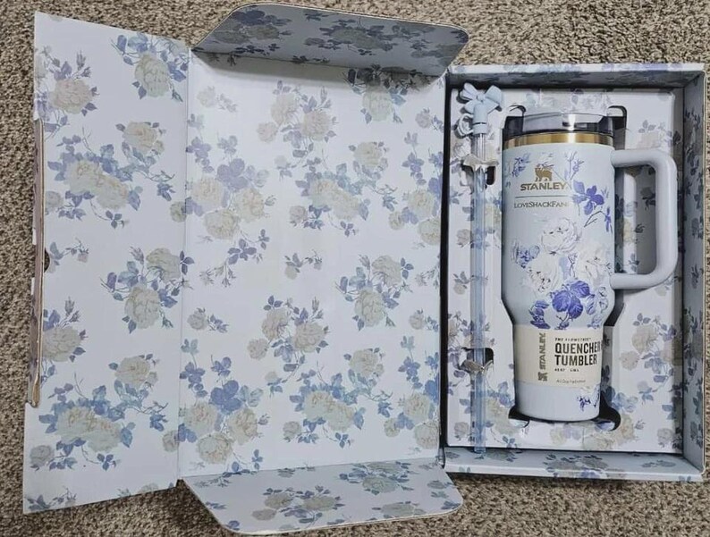May include: A white Stanley Quencher tumbler with a handle, floral design, and a straw, presented in a matching floral-patterned box. The tumbler has the text "Stanley Loveshackfancy" and "Quencher Tumbler".