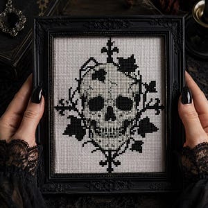 May include: A black and white cross-stitch embroidery featuring a detailed human skull surrounded by ivy leaves and decorative cross motifs. The piece is encased in a rectangular black frame with ornate detailing along the edges.