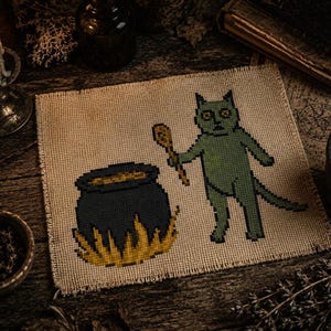 May include: Cross-stitch art featuring a green cat holding a wooden spoon, standing next to a black cauldron with yellow flames. The artwork is on a tan fabric, with a rustic, witchy theme. A lit candle and old book are in the background.