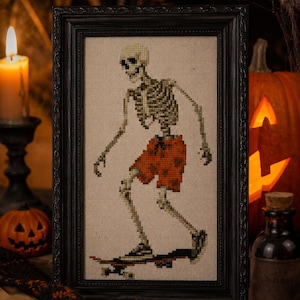 May include: Framed cross-stitch art of a skeleton skateboarding, wearing orange shorts. The skeleton is in a black frame, with a Halloween theme. A lit candle, carved pumpkin, and potion bottle are in the background.