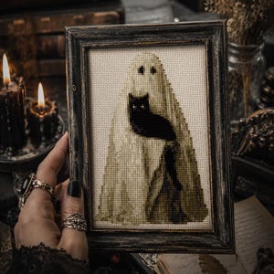 May include: Framed cross-stitch art featuring a ghost and a black cat. The ghost is draped in a cream-colored sheet, and the cat is perched on the ghost's form. The frame is dark wood, and the artwork has a vintage, gothic aesthetic.
