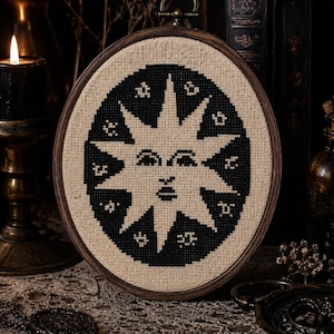 Può includere: Un telaio da ricamo circolare contenente un disegno a punto croce su tessuto beige. Il design presenta uno sfondo ovale nero con un motivo solare centrale in beige, che mostra un volto umano con gli occhi chiusi e piccoli simboli a stella.