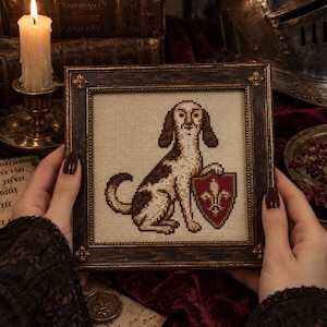 May include: A square cross-stitch embroidery in a dark, distressed wooden frame. The needlework depicts a brown and white dog sitting upright, holding a red shield featuring a gold fleur-de-lis emblem against a cream-colored fabric background.