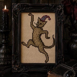 May include: A cross-stitch embroidery of a dancing cat in a tan shade, wearing a small blue and red patterned pointed hat. The artwork is displayed within a dark, ornate, rectangular frame with intricate carved details along the edges.