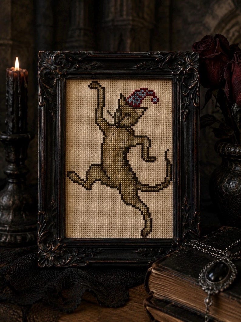 May include: A cross-stitch embroidery of a dancing cat in a tan shade, wearing a small blue and red patterned pointed hat. The artwork is displayed within a dark, ornate, rectangular frame with intricate carved details along the edges.