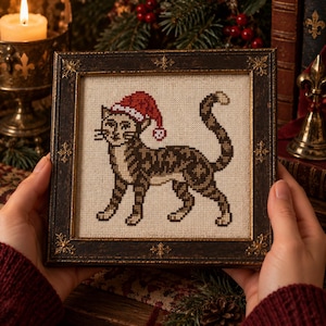 May include: A cross-stitch embroidery on light beige fabric featuring a brown tabby cat wearing a red and white Santa hat. The embroidery is encased in a dark, ornate, rectangular wooden frame with gold-toned decorative corner accents.