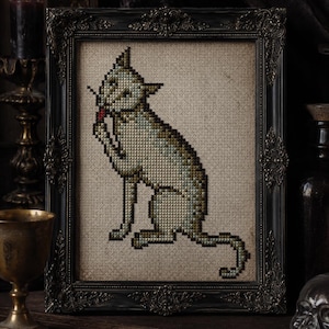 May include: A cross-stitch embroidery of a seated cat grooming itself, rendered in muted green, brown, and gray tones on a beige fabric background. The piece is housed in an ornate, dark, rectangular frame with intricate carved detailing.