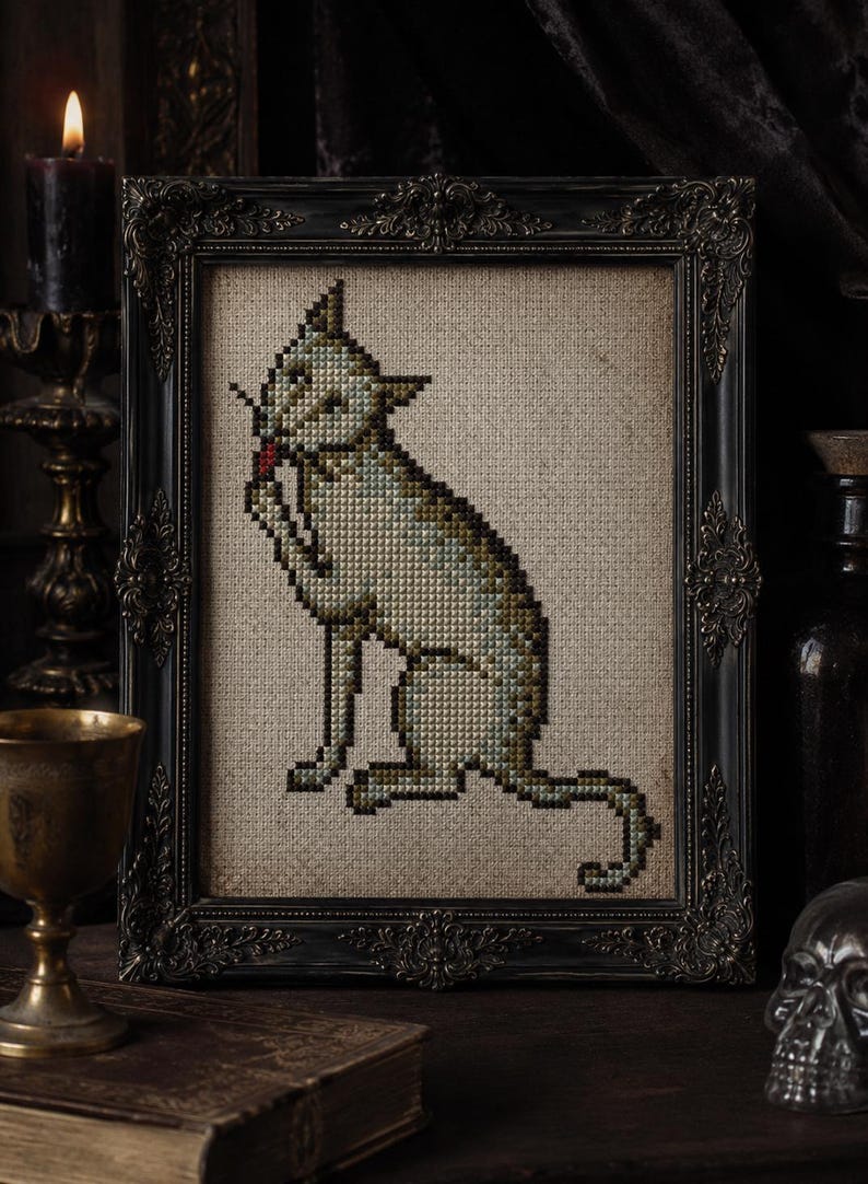 May include: A cross-stitch embroidery of a seated cat grooming itself, rendered in muted green, brown, and gray tones on a beige fabric background. The piece is housed in an ornate, dark, rectangular frame with intricate carved detailing.