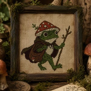Könnte beinhalten: Eine Kreuzstichstickerei auf beigem Stoff zeigt einen grünen Frosch mit einem rot-weiß gepunkteten Pilzhut und einem dunkelbraunen Umhang. Der Frosch trägt eine braune Umhängetasche und hält einen dünnen Wanderstock, gerahmt in dunklem Holz.