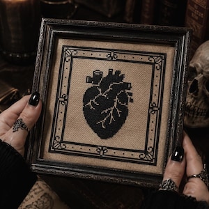 May include: A framed cross-stitch featuring a black anatomical heart design. The artwork is set against a tan fabric background and framed in a dark, ornate frame. The piece is held by hands with black nail polish and gothic rings.