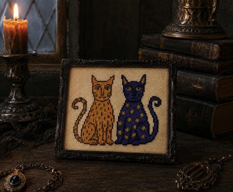 May include: A framed cross-stitch embroidery featuring two stylized cats sitting side by side. One cat is orange with darker orange spots, and the other is dark blue with yellow star-shaped patterns, both set against a light beige fabric background.