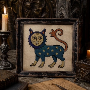 May include: A square framed cross-stitch embroidery featuring a stylized cat with a blue body, yellow star patterns, and a long orange tail. The cat has a human-like face surrounded by a dark blue mane, all set against a natural beige fabric background.