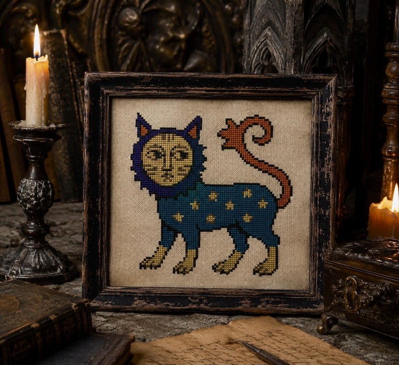 May include: A square framed cross-stitch embroidery featuring a stylized cat with a blue body, yellow star patterns, and a long orange tail. The cat has a human-like face surrounded by a dark blue mane, all set against a natural beige fabric background.