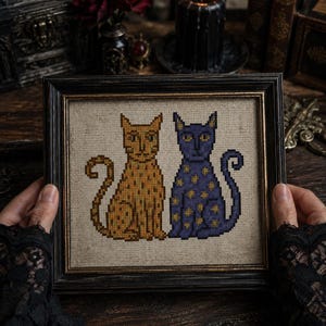 May include: A framed cross-stitch embroidery featuring two stylised cats sitting side by side. One cat is golden yellow with dark speckles, and the other is deep purple with yellow star-like spots. The piece is set in a dark, ornate wooden frame.