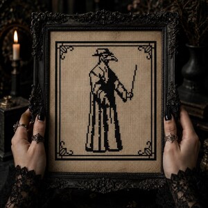 May include: A black and beige cross-stitch embroidery featuring a plague doctor figure in a long coat and wide-brimmed hat, holding a thin cane. The design is enclosed in a black geometric border and housed within an ornate black rectangular frame.