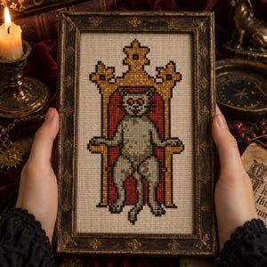 May include: A cross-stitch embroidery in a dark, ornate rectangular frame. The design features a light-coloured cat sitting on a tall, golden throne with a deep red background. The cat has large, light-coloured eyes and is centred within the frame.