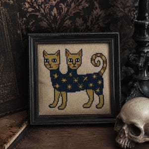 May include: A framed cross-stitch embroidery featuring a two-headed cat with yellow fur and blue eyes. The cat has a dark blue body patterned with yellow stars and a curled tail, all stitched onto a light tan fabric within a black frame.