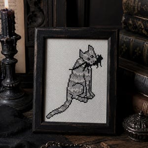 May include: A framed cross-stitch embroidery featuring a pixelated gray cat with a black mouse in its mouth. The cat is depicted in a seated position with a long tail extending outward, rendered in shades of gray on a light-colored fabric background.
