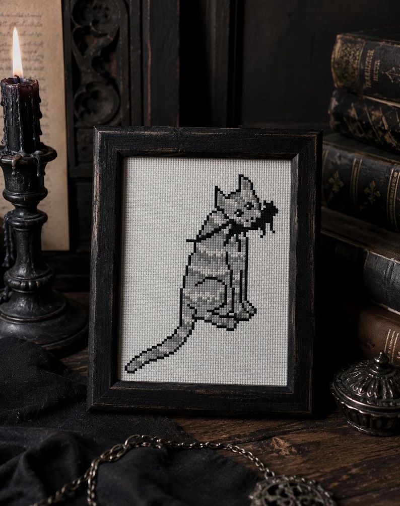 May include: A framed cross-stitch embroidery featuring a pixelated gray cat with a black mouse in its mouth. The cat is depicted in a seated position with a long tail extending outward, rendered in shades of gray on a light-colored fabric background.