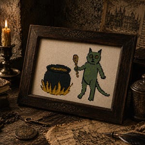 May include: A framed cross-stitch embroidery featuring a green cat-like creature holding a wooden spoon next to a dark blue cauldron with yellow flames at the base, all set against a light-colored fabric background within a dark wooden frame.