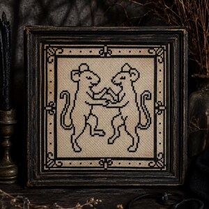 May include: A framed cross-stitch featuring two stylized black mice holding hands. The artwork is set against a beige background and framed in a distressed black wooden frame. The piece has a vintage, gothic aesthetic.