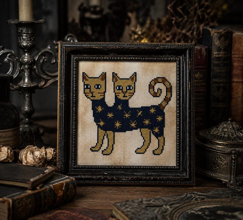 May include: A framed cross-stitch embroidery featuring a two-headed cat with tan fur and blue eyes. The cat wears a dark blue garment decorated with gold star patterns. The embroidery is set against a beige fabric background in a dark wooden frame.