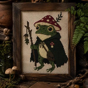 Puede incluir: Arte de punto de cruz de una rana verde con gorro y capa de hongo, sosteniendo una linterna y un bastón. La obra de arte enmarcada tiene un marco de madera rústico. La rana está sobre un fondo neutro, con elementos forestales en primer plano.