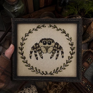 May include: A square framed cross-stitch embroidery featuring a pixelated jumping spider in shades of brown, tan, and black, centred within a circular wreath of green leaves on a light beige fabric background, enclosed in a dark ornate frame.