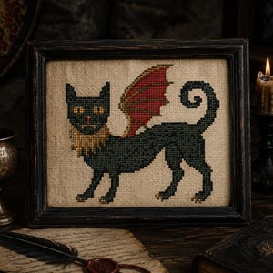 May include: Cross-stitch art of a black cat-like creature with red and gold wings, framed in a dark wooden frame. The creature has a gold mane and yellow eyes. The background is a neutral tan fabric.