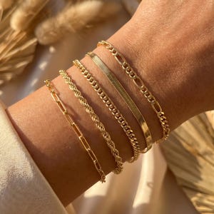 May include: A collection of gold bracelets in various styles, including a chain link, rope, and herringbone design. The bracelets are stacked on a wrist, showcasing their different textures and shine. The warm lighting highlights the gold color.