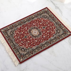 Op de afbeelding: Een rechthoekig, sierlijk tapijt met een dieprode basis en ingewikkelde bloempatronen. Het tapijt heeft een centraal medaillon en een rand met zwarte, beige en grijze accenten. Crèmekleurige franjes omzomen de randen.