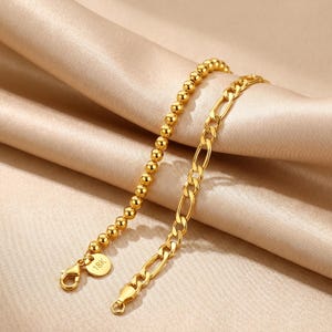 May include: Two gold-colored bracelets. One bracelet features small, round beads, while the other has a chain link design. Both bracelets have a clasp closure. A small, round charm with the text "18K" is attached to the beaded bracelet.