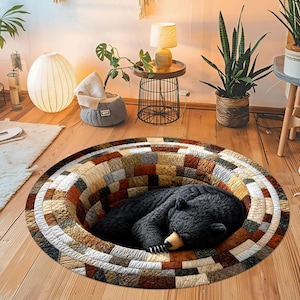 Op de afbeelding: Een rond vloerkleed met een 3D-optische illusie van een diep, cirkelvormig gat met een getextureerde, veelkleurige patchworkrand in bruin-, beige- en grijstinten. Een zwarte beer slaapt in het midden van het gat.