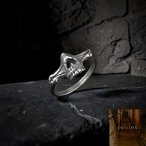 Puede incluir: Un anillo plateado con un diseño de calavera. La calavera presenta cuencas de los ojos, una cavidad nasal y dientes. El anillo se muestra sobre un fondo oscuro y texturizado, resaltando los intrincados detalles de la calavera.