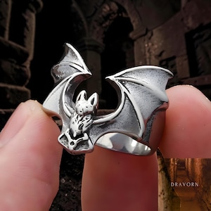 May include: A silver-colored ring featuring a detailed bat design. The bat has outstretched wings and is centered on the ring's face. The ring is held up by a finger, showcasing the intricate details of the bat's features.