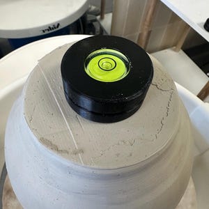 May include: A circular black spirit level with a bright neon yellow liquid center and a centered bubble, resting on top of a large, unfinished, light gray clay vessel that is currently being shaped on a pottery wheel in a studio setting.