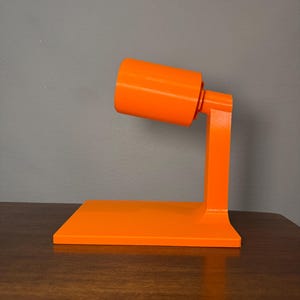 May include: A bright orange desk lamp base featuring a flat rectangular platform, a vertical support arm, and a cylindrical socket head. The structure is made of a smooth, solid-colored material with a glossy finish and a minimalist geometric design.