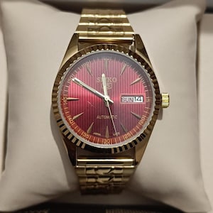 Op de afbeelding: Een goudkleurig Seiko 5 automatisch polshorloge met een levendige rode sunburst wijzerplaat met verticale krijtstrepen, goudkleurige wijzers en uurmarkeringen, een geribbelde lunette en een dag-datumvenster op de drie-uurpositie aan een schakelarmband.