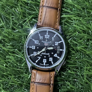 May include: A Seiko 5 automatic wristwatch with a silver-tone stainless steel case and a brown leather strap featuring a crocodile-embossed texture. The black dial displays white Arabic numerals, a date window, and a textured geometric pattern.