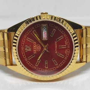 Op de afbeelding: Een goudkleurig Seiko 5 automatisch polshorloge met een dieprode ronde wijzerplaat, goudkleurige uurmarkeringen, een geribbelde rand en een bijpassende goudkleurige metalen schakelarmband. De wijzerplaat toont de tekst Seiko 5, Automatic en Japan Made.