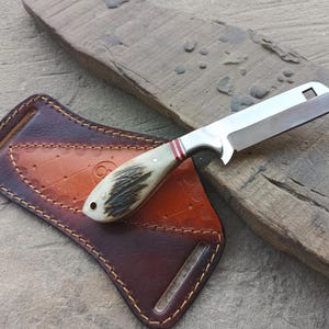 May include: A fixed-blade knife with a rectangular steel blade featuring a square cutout near the tip, a handle made of textured antler with red and white accents, and a brown leather sheath with decorative stitching and a belt loop attachment.