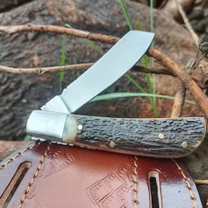May include: A folding pocket knife with a single silver-toned blade and a textured, dark brown handle featuring three small brass pins. The knife is positioned resting on a smooth, dark brown leather sheath with decorative embossed patterns.