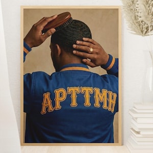 May include: A framed print featuring a person brushing their hair. The person is wearing a blue jacket with gold lettering that reads "APTTHM". The jacket has gold and blue striped cuffs and collar. The background is a neutral beige.